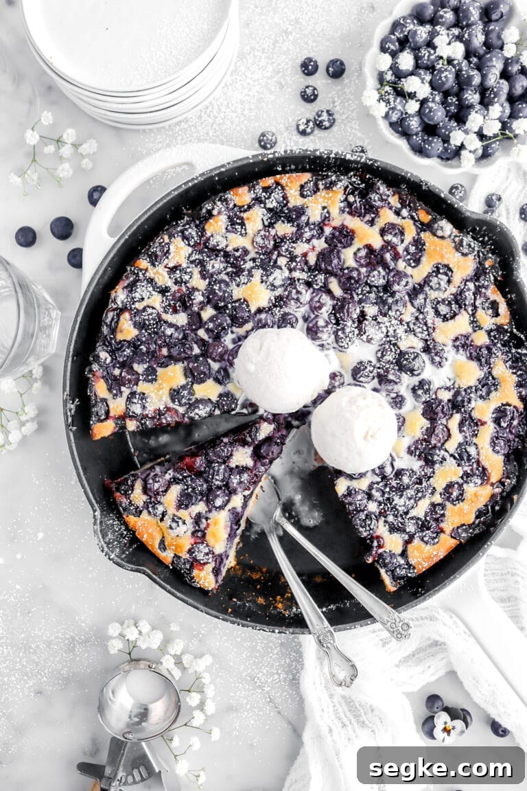 cropped close up of blueberry skillet with slices cut out of it and two forks resting in skillet with two scoops of ice cream on top of cobbler, white flowers and blueberries around on marble surface.