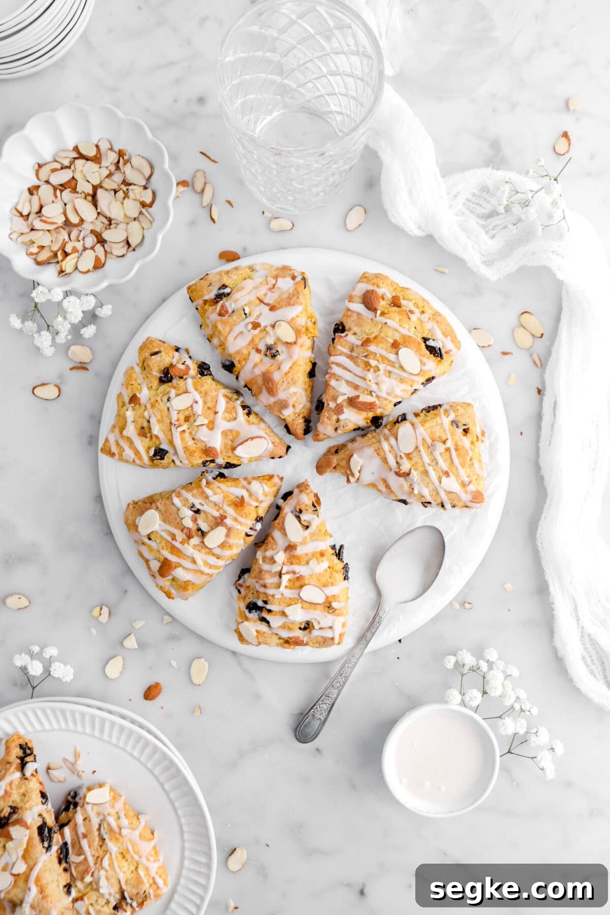 overhead shot of cherry almond scones on upside down white plate with white flowers and almond around on marble surface.