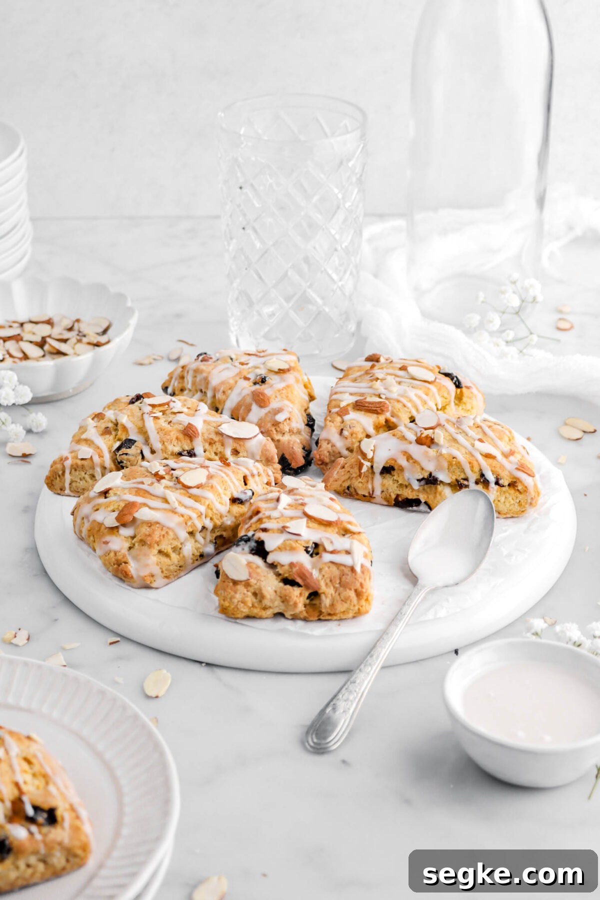 front shot of scones on upside down white plate with spoon beside, empty glasses, and bowl of alices almonds behind.