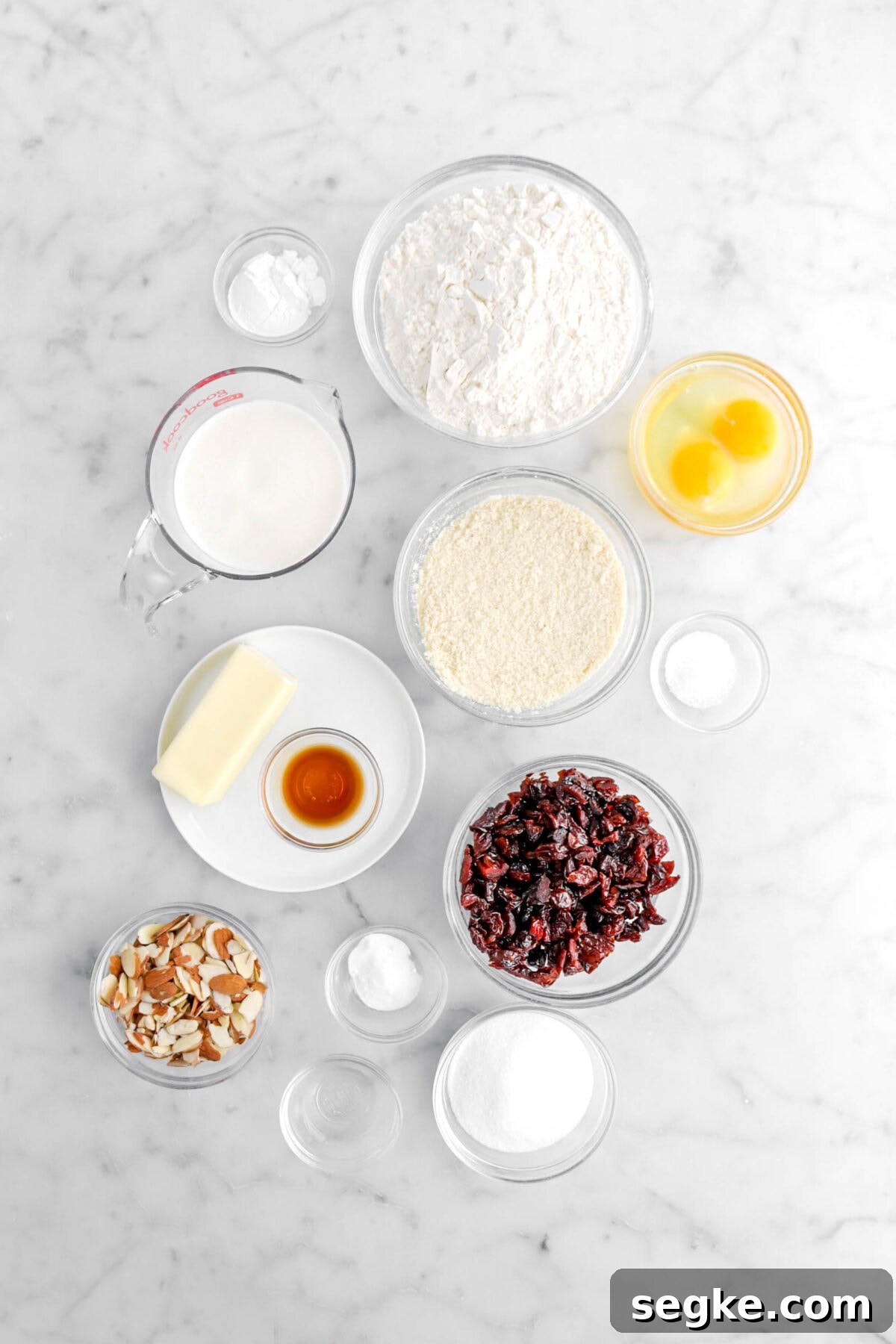 baking powder, flour, milk, eggs, almond flour, butter, vanilla, dried cherries, salt, sliced almonds, baking soda, almond extract, and sugar on marble surface.