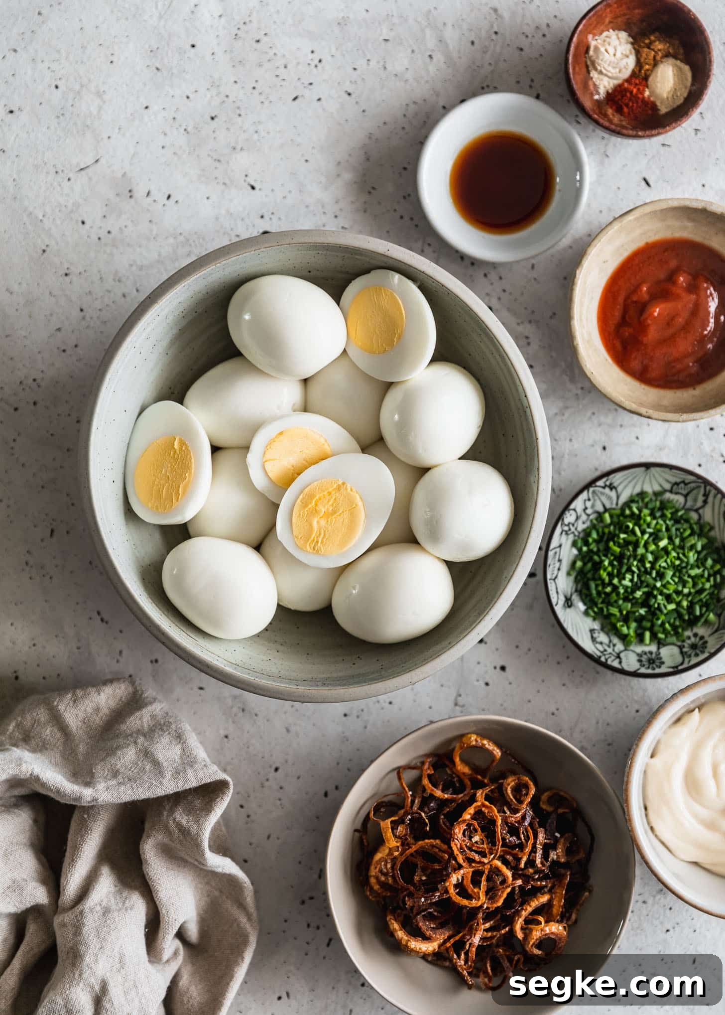White and grey bowls with hard boiled eggs, Sriracha, mayo, and spices on a grey counter next to a beige linen.