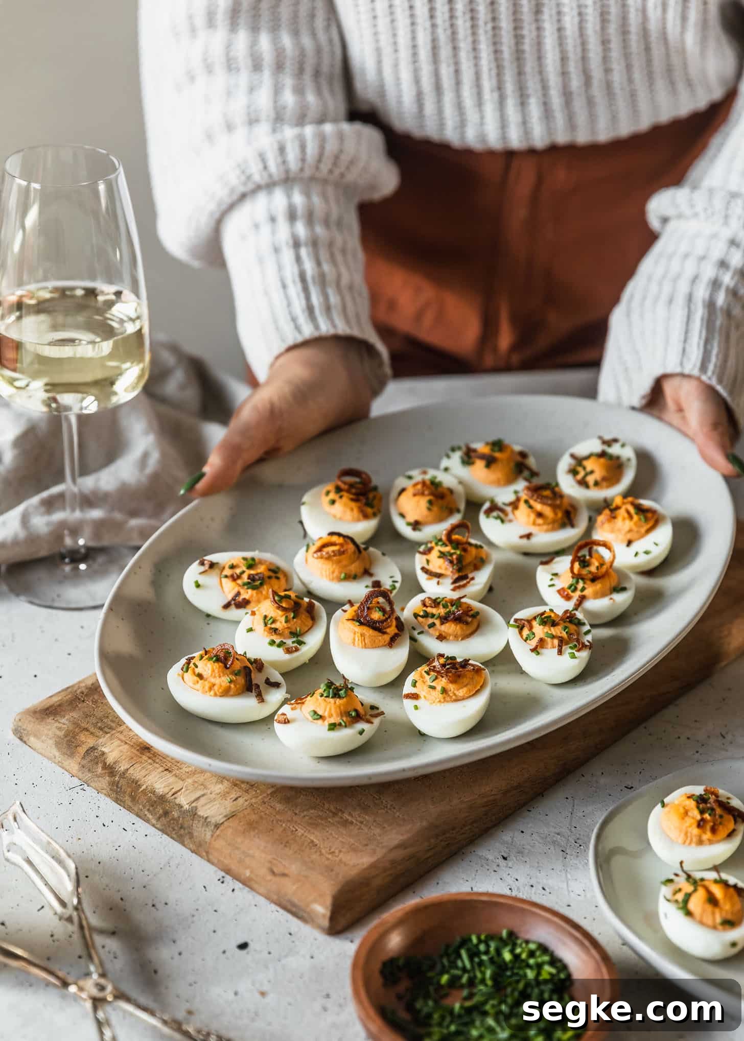 A woman in a white sweater and orange pants placing a white tray of spicy deviled eggs on a white counter next to a wood bowl of chives and glasses of white wine.