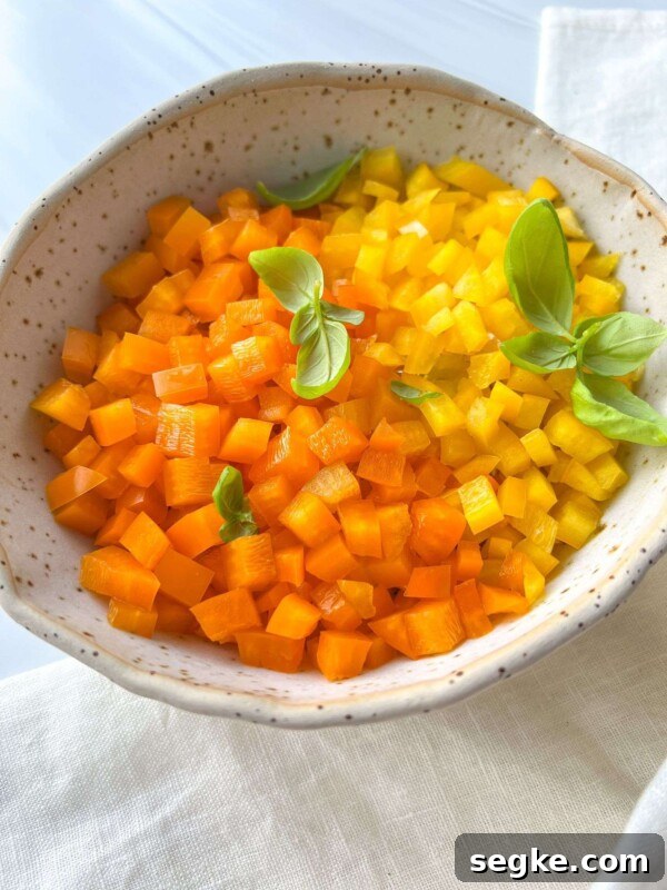 A bowl filled with small, neatly diced orange and yellow bell peppers, garnished with fresh basil leaves, sits on a light-colored cloth—perfect for showcasing your skills in How To Dice Bell Peppers.