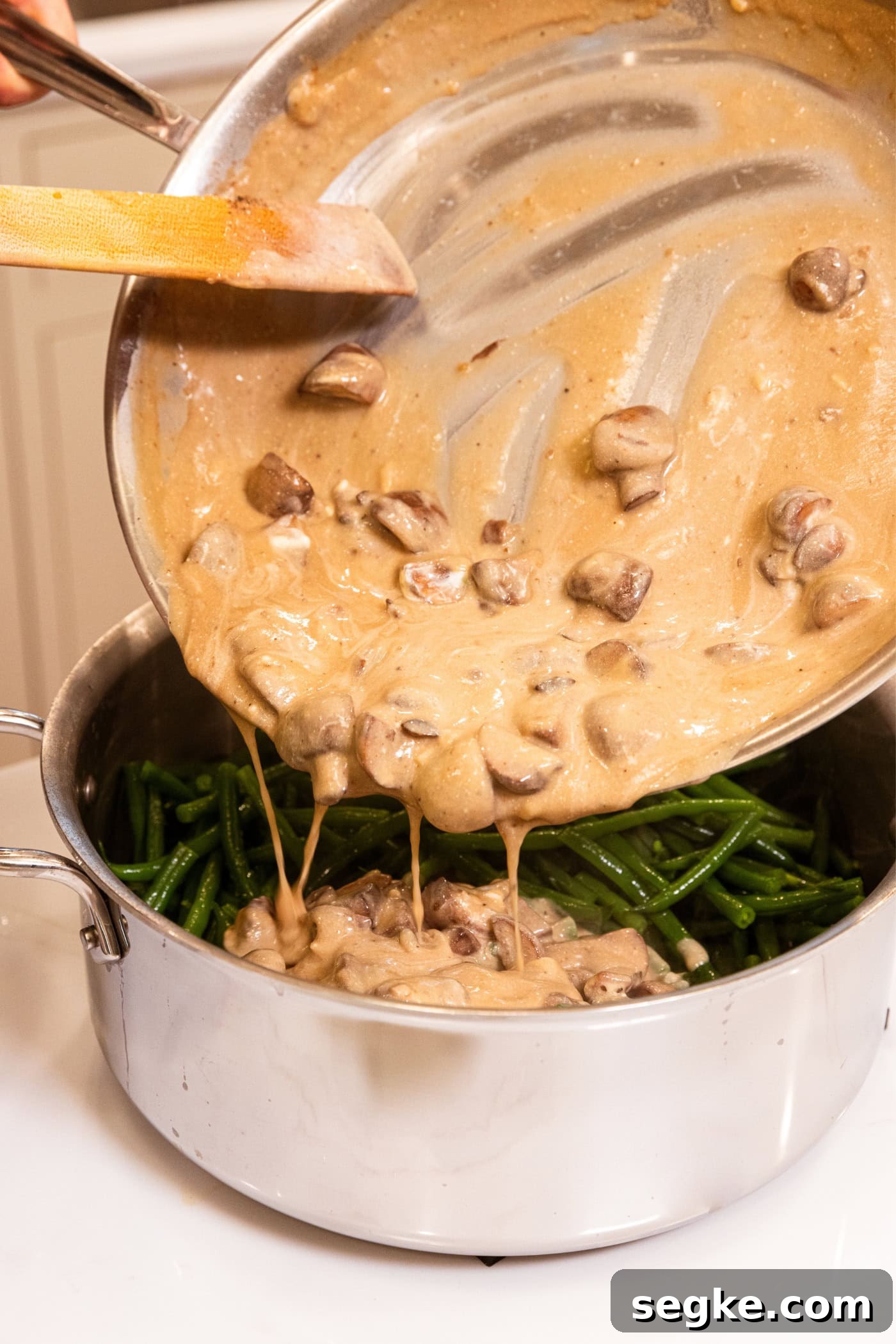 Creamy Homemade Green Bean Casserole Recipe 10 Green beans in a large stainless steel pot with a mushroom sauce being poured over the top.