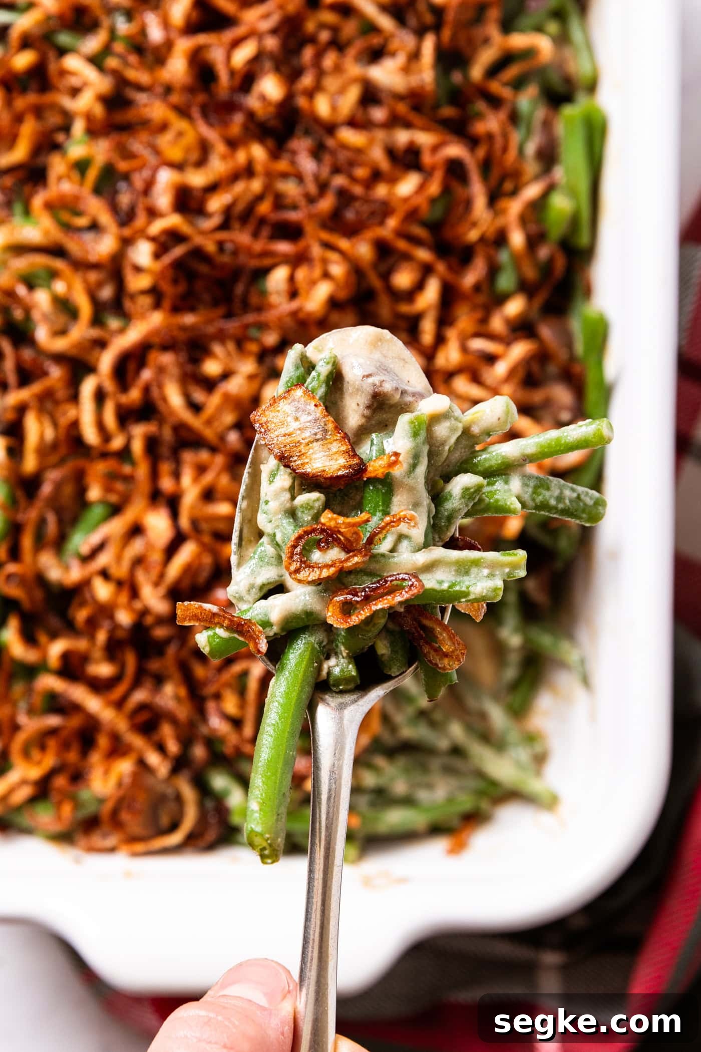Creamy Homemade Green Bean Casserole Recipe 3 White baking dish filled with green beans with mushroom sauce with crispy fried shallots on top with hand holding spoonful after scooping from baking dish with Christmas napkin in front.