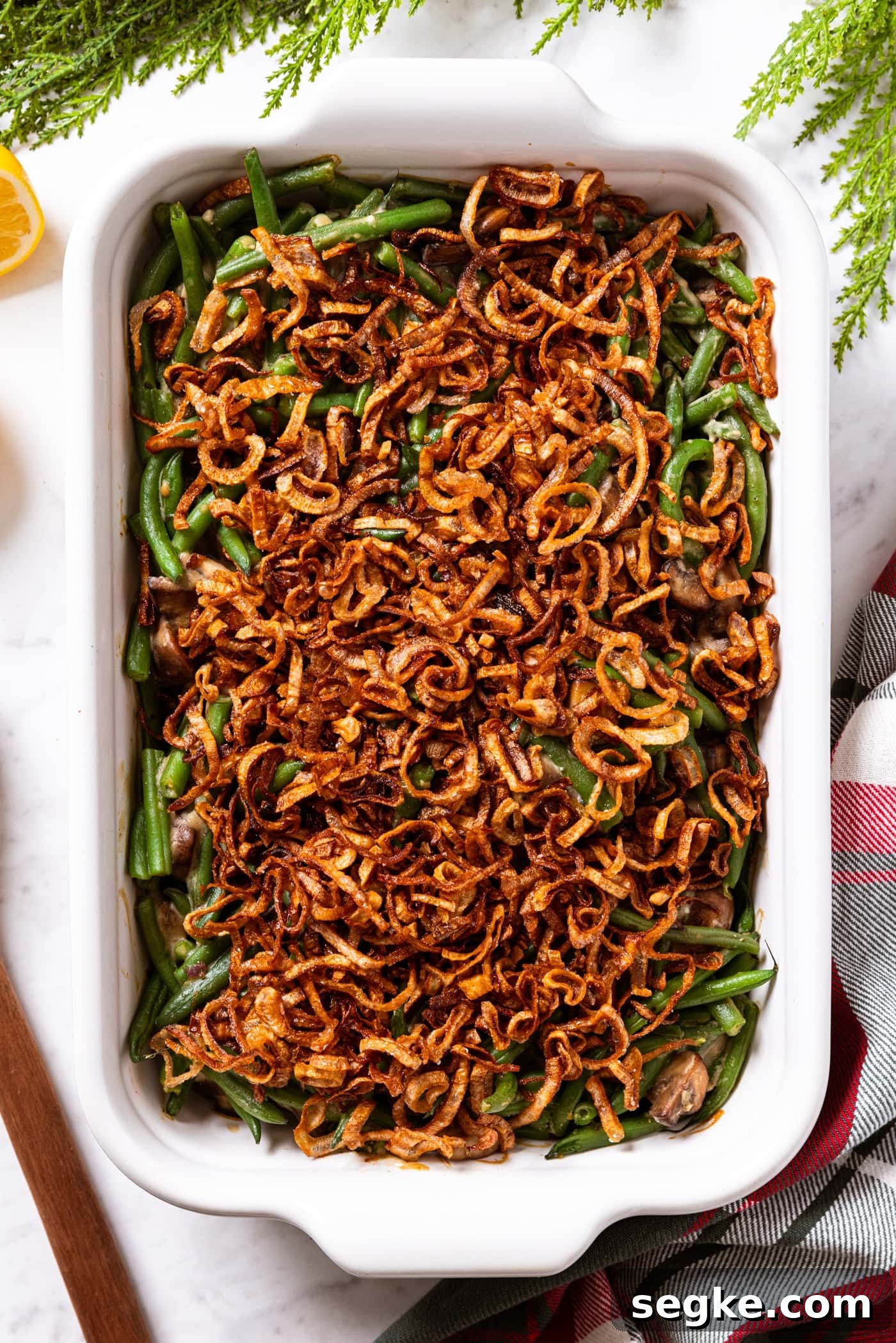 Creamy Homemade Green Bean Casserole Recipe 13 White baking dish filled with baked green bean casserole sitting on white marble surface with greenery all around as well as Christmas napkin topped with crispy fried shallots.