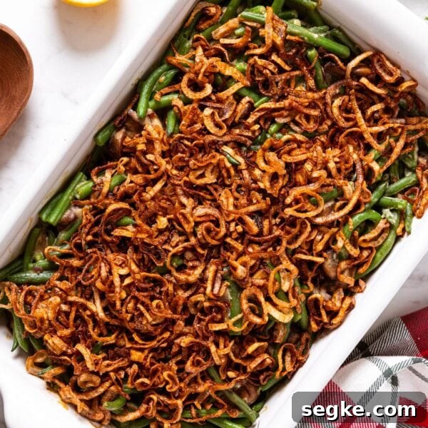 White baking dish filled with baked green bean casserole topped with crispy fried scallions with wooden spoon, lemon, and Christmas napkin to the side all on marble surface.