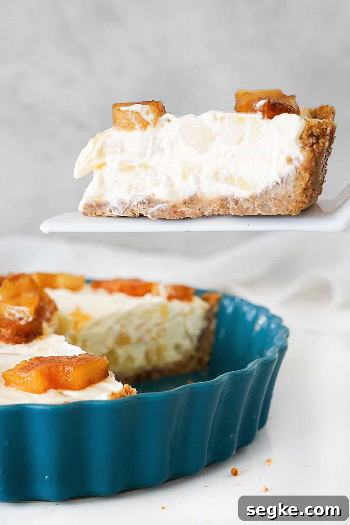 No-Bake Pineapple Cream Cheese Pie 12 Slice of pineapple cheese pie on a white cake server. Teal dish with pie below. Light grey background.