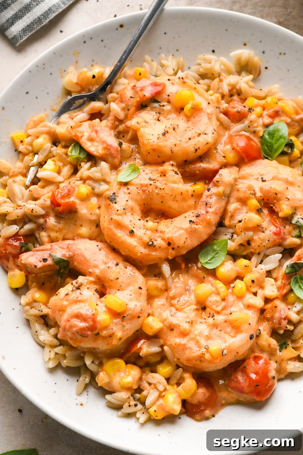 A plate of creamy shrimp with corn, tomatoes, and herbs served over orzo pasta, garnished with fresh basil and black pepper. A fork rests on the edge of the dish.