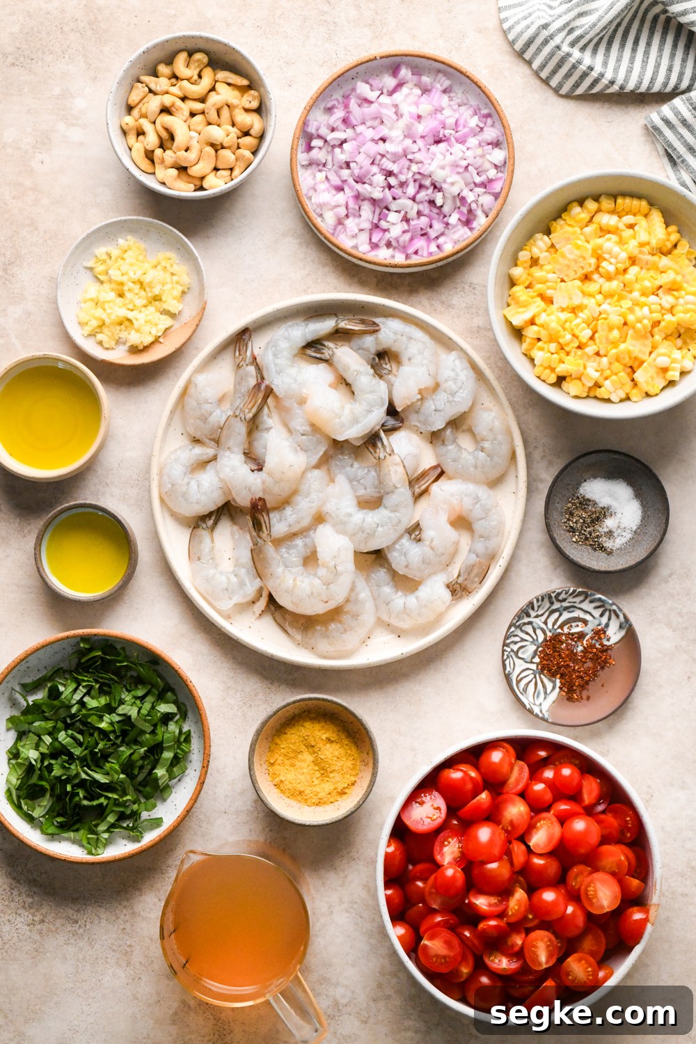 Overhead view of raw shrimp on a plate, surrounded by bowls of chopped cherry tomatoes, corn, raw cashews, olive oil, fresh basil, diced shallots, minced garlic, and a cup of broth on a light surface.