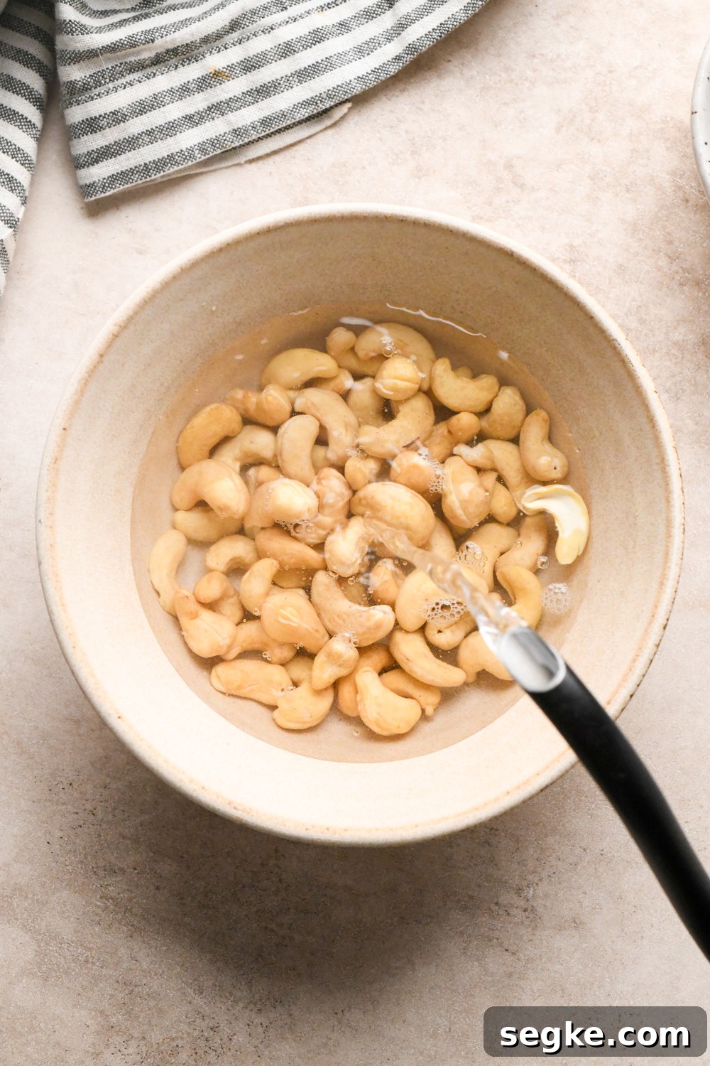 A bowl of raw cashews soaking in water, with a stream of water being poured in, on a light countertop next to a striped cloth.