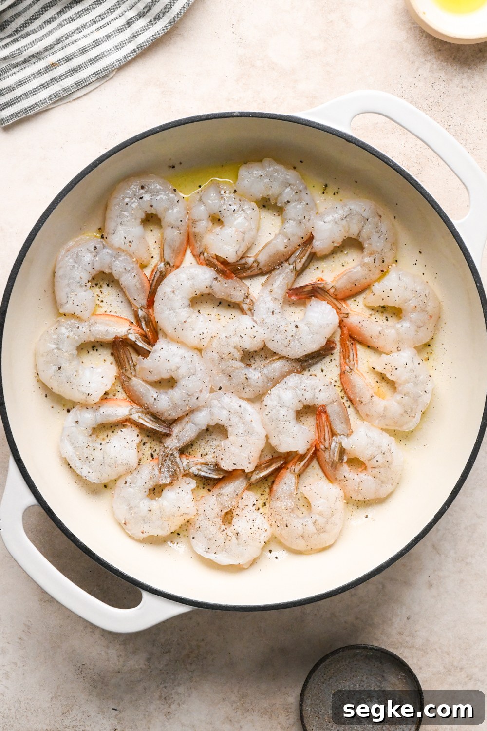 Raw shrimp arranged in a circular pattern in a white skillet with olive oil, seasoned with salt and pepper, cooking on one side.