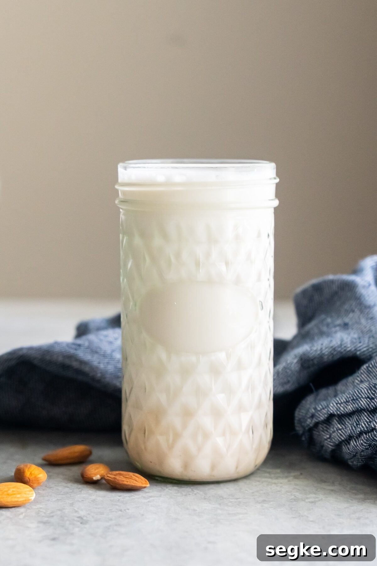 A glass jar filled with almond milk sits on a countertop beside a few almonds and a gray cloth.