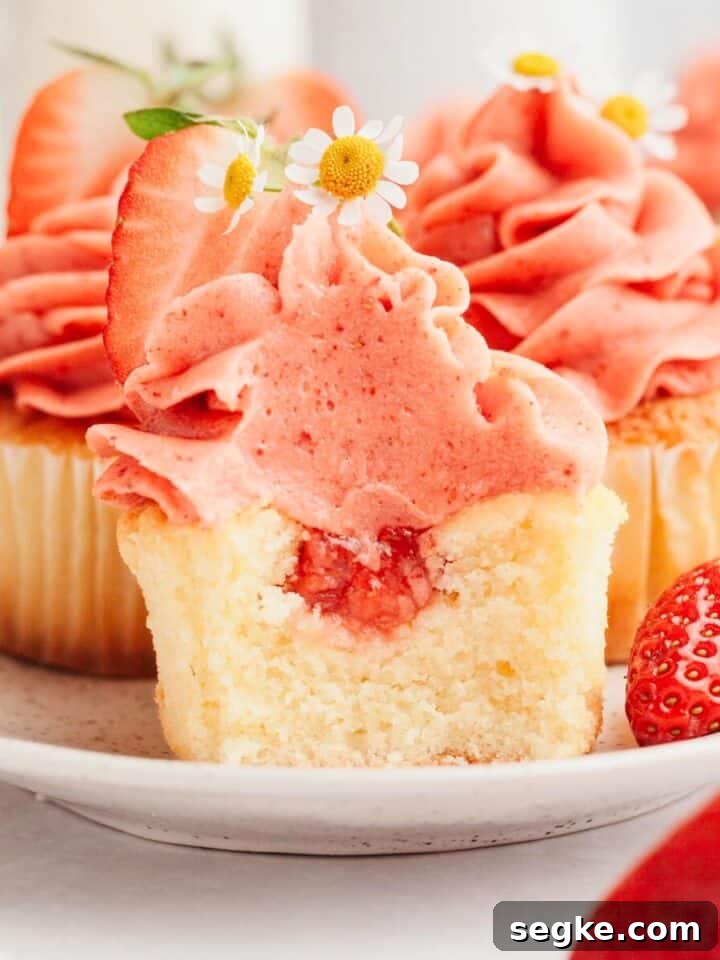 A vanilla cupcake with strawberry filling and strawberry frosting, a bite has been taken out of it.