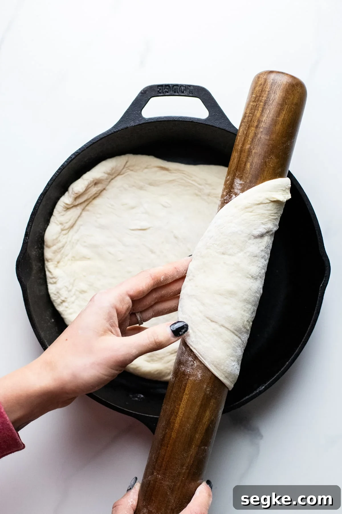 How to Make Crispy Cast Iron Skillet Pizza 3 hands using rolling pin to transfer pizza dough into cast iron skillet on white background.
