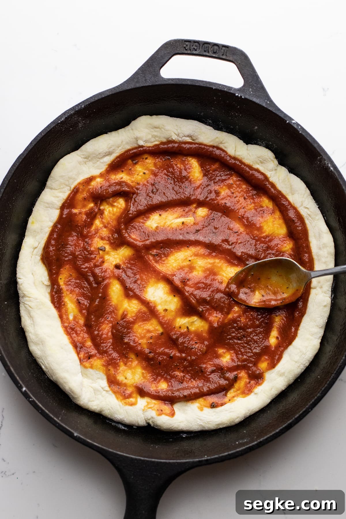 How to Make Crispy Cast Iron Skillet Pizza 5 A spoon smoothing out tomato sauce on pizza dough in a cast iron skillet.