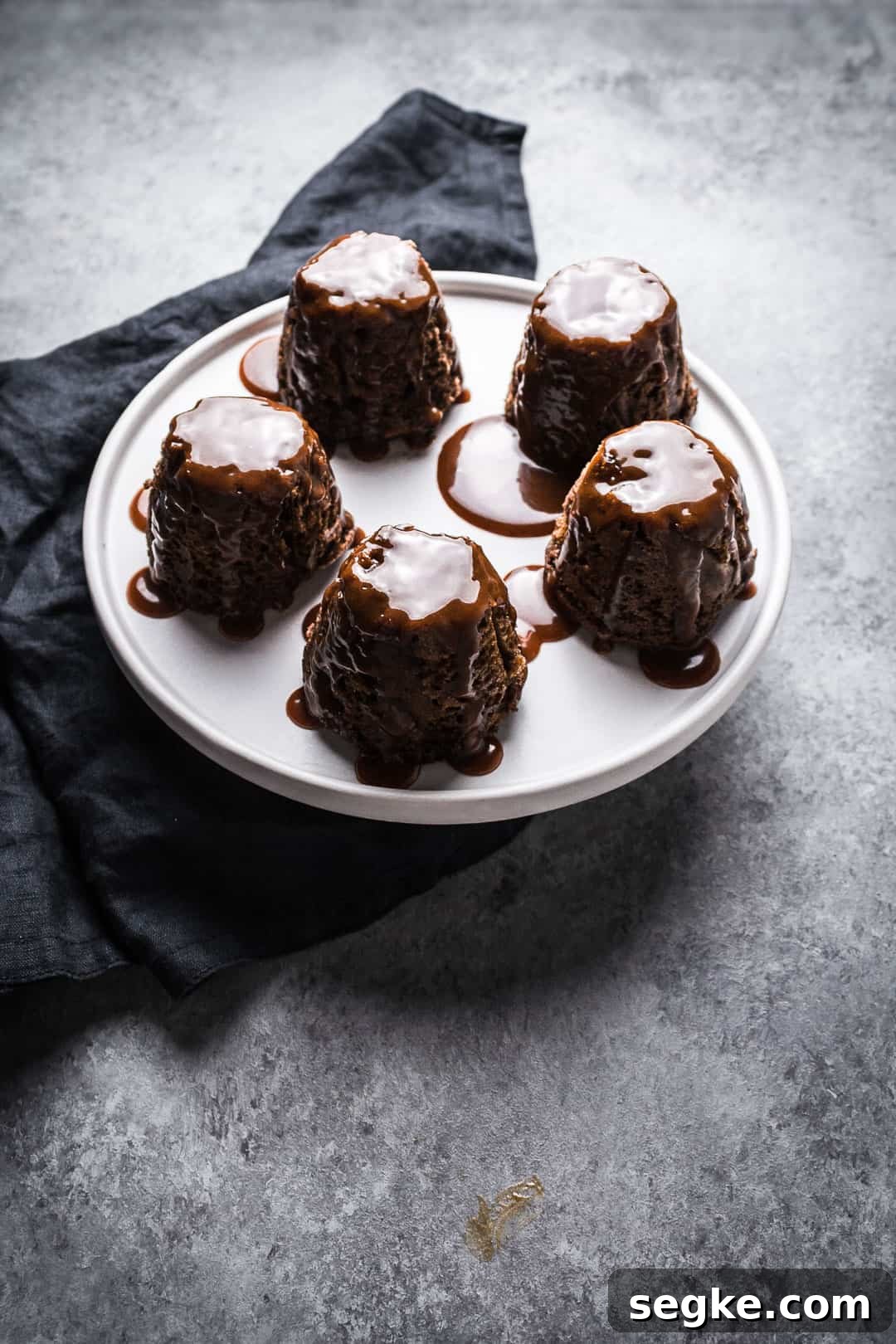 Forty five degree angle view of five backlit apple ginger mini cakes topped with salted caramel sauce on a white plate with a grey background