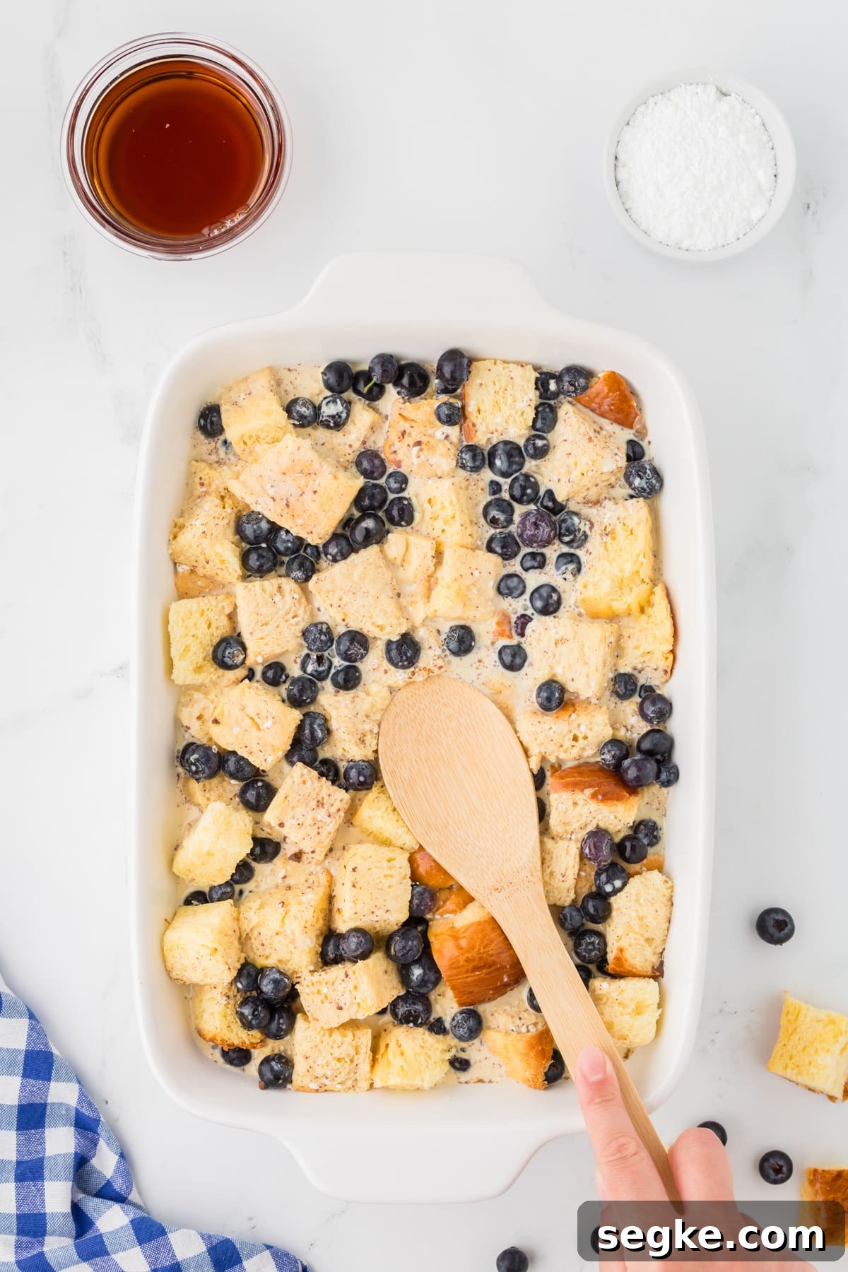 Using a wooden spoon to press bread cubes into a custard mixture in a baking dish.