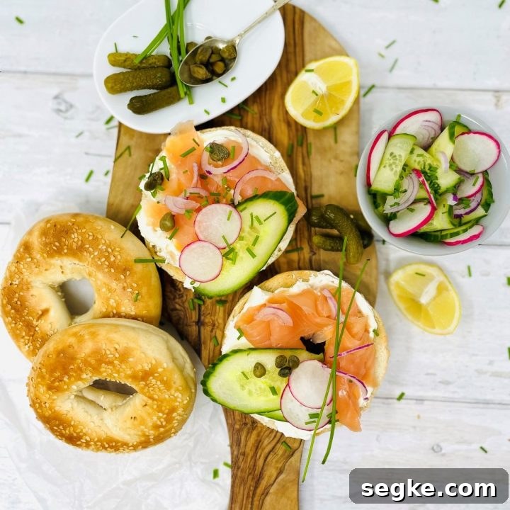 Image shows a wooden chopping board with a bagel sliced in half topped with smoked salmon, chives, capers, radish, onion and cucumber, there is also a bowl of cucumber,radish and onions as well as a plate with mini pickles and gherkins on, there is also two bagels and a sliced lemon next to the chopping board