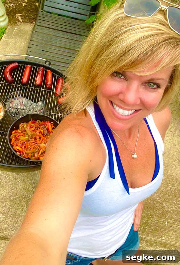 Grilled Sausage with Peppers and Onions Recipe 3 Jennifer with weber grill behind grilling sausages, onions, and peppers