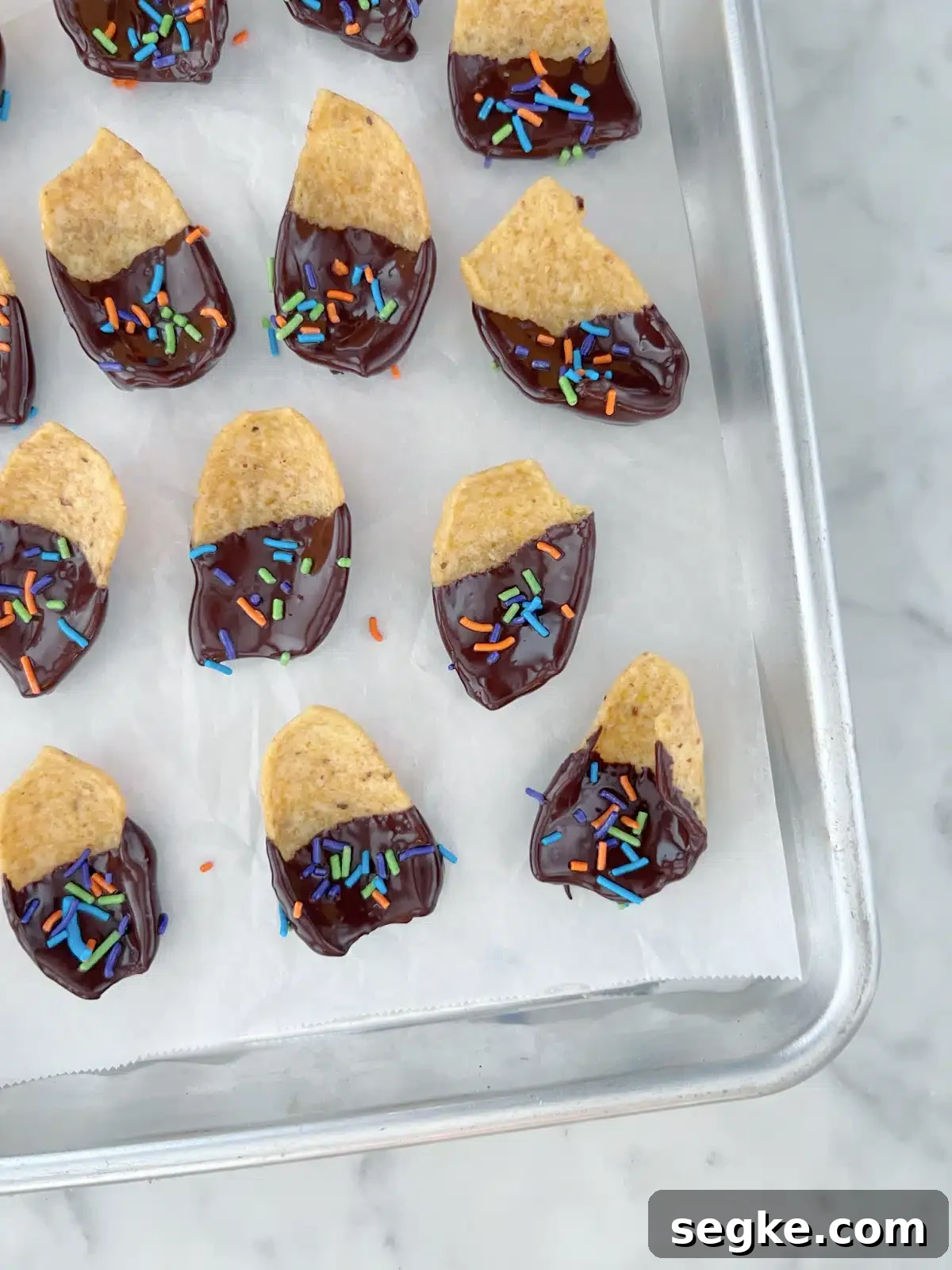 Overhead view of a parchment lined baking pan of chocolate dipped corn chips covered in sprinkles. The chocolate is shiny from being freshly dipped and has not yet dried.