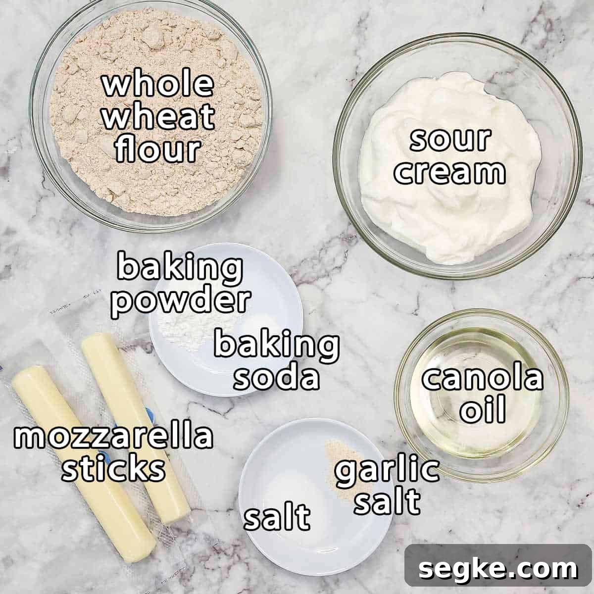 Flaky Mozzarella-Stuffed Whole-Wheat Biscuits Recipe 3 Overhead shot of ingredients - whole wheat flour, sour cream, baking powder, baking soda, canola oil, mozzarella sticks, salt and garlic salt.