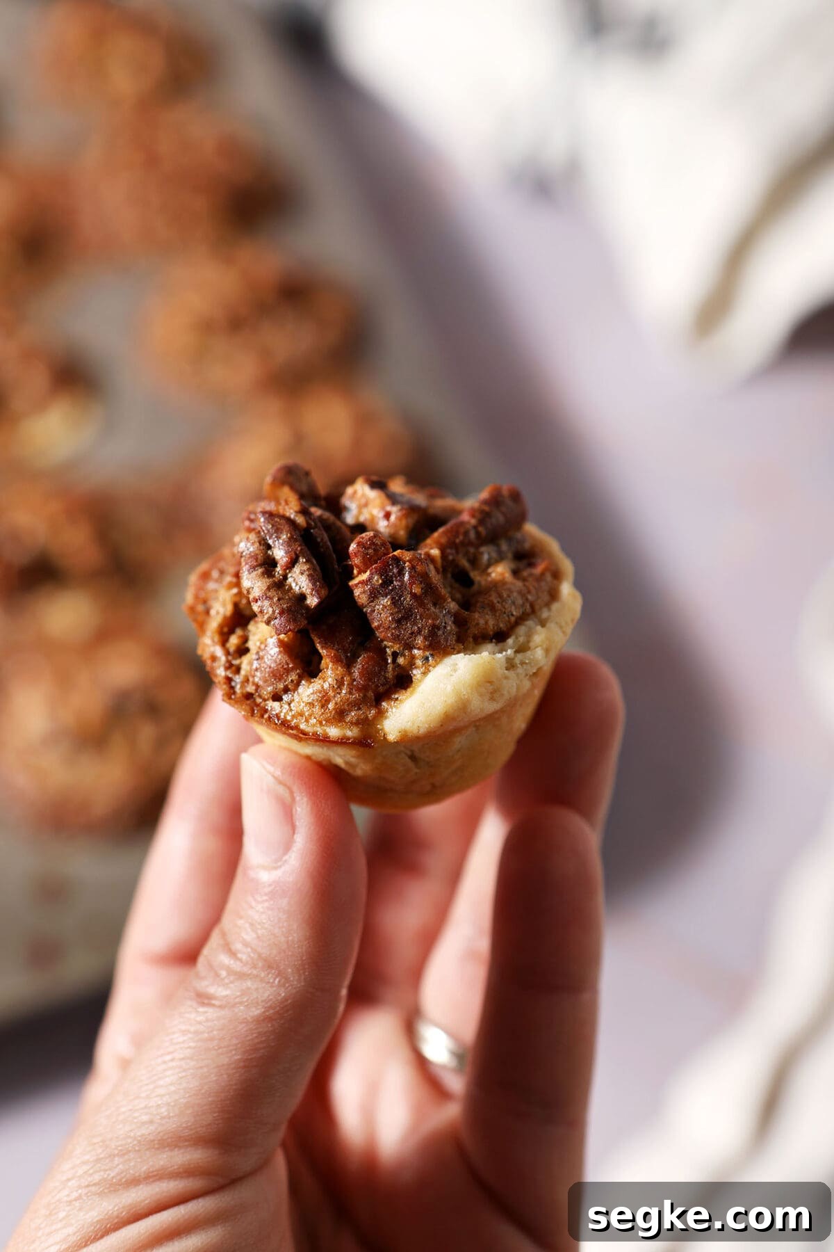 a hand holds a pecan tassie above a mini muffin tin of more tassies on a purple surface