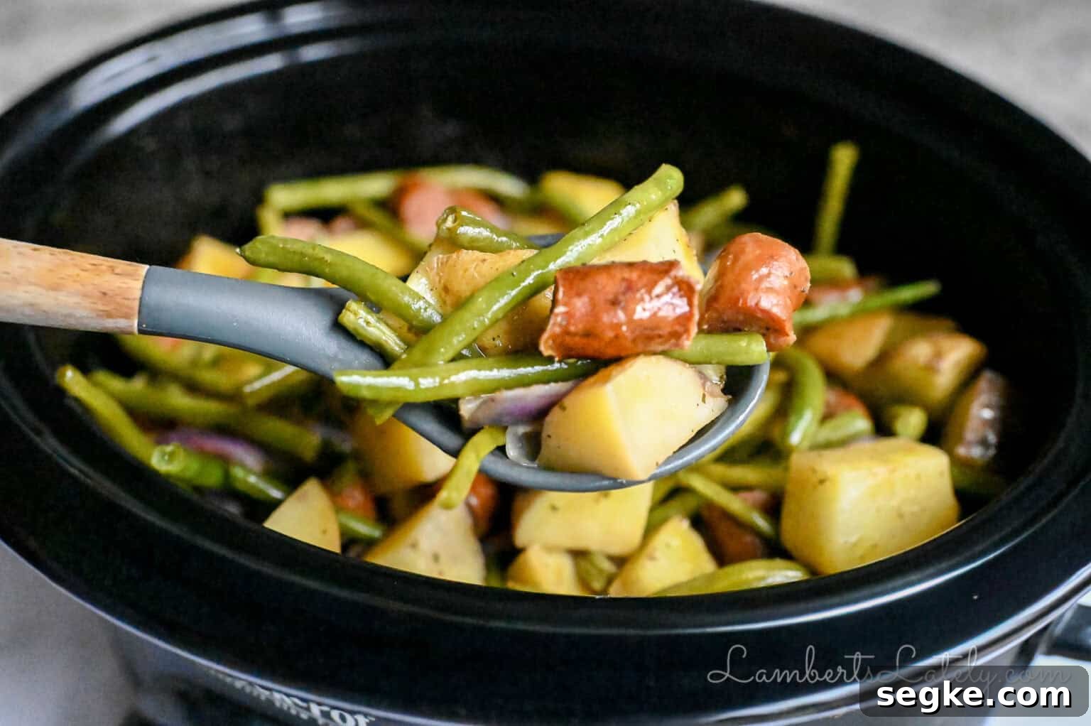 Slow Cooker Southern Swamp Potatoes Recipe 8 holding a spoonful of swamp potatoes with green beans and sausage in front of a crock pot.