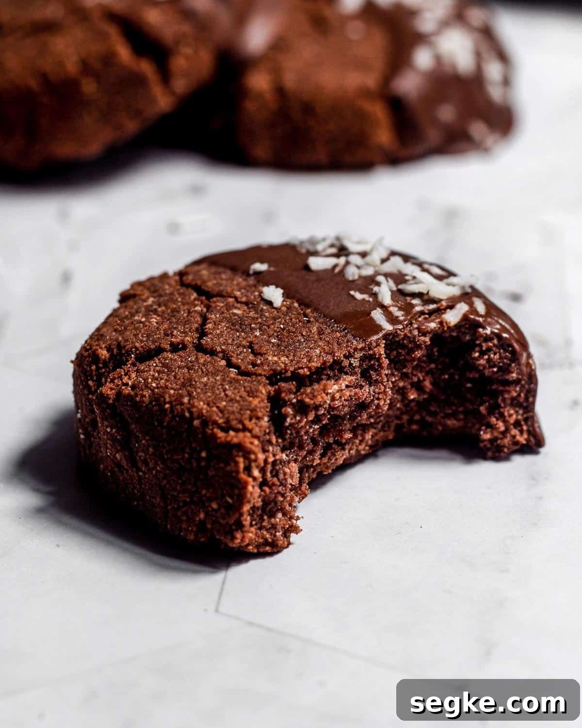 Gluten-Free Chocolate Coconut Biscuits Recipe 4 A chocolate coconut biscuit with a bite missing. There are two more cookies in the background.