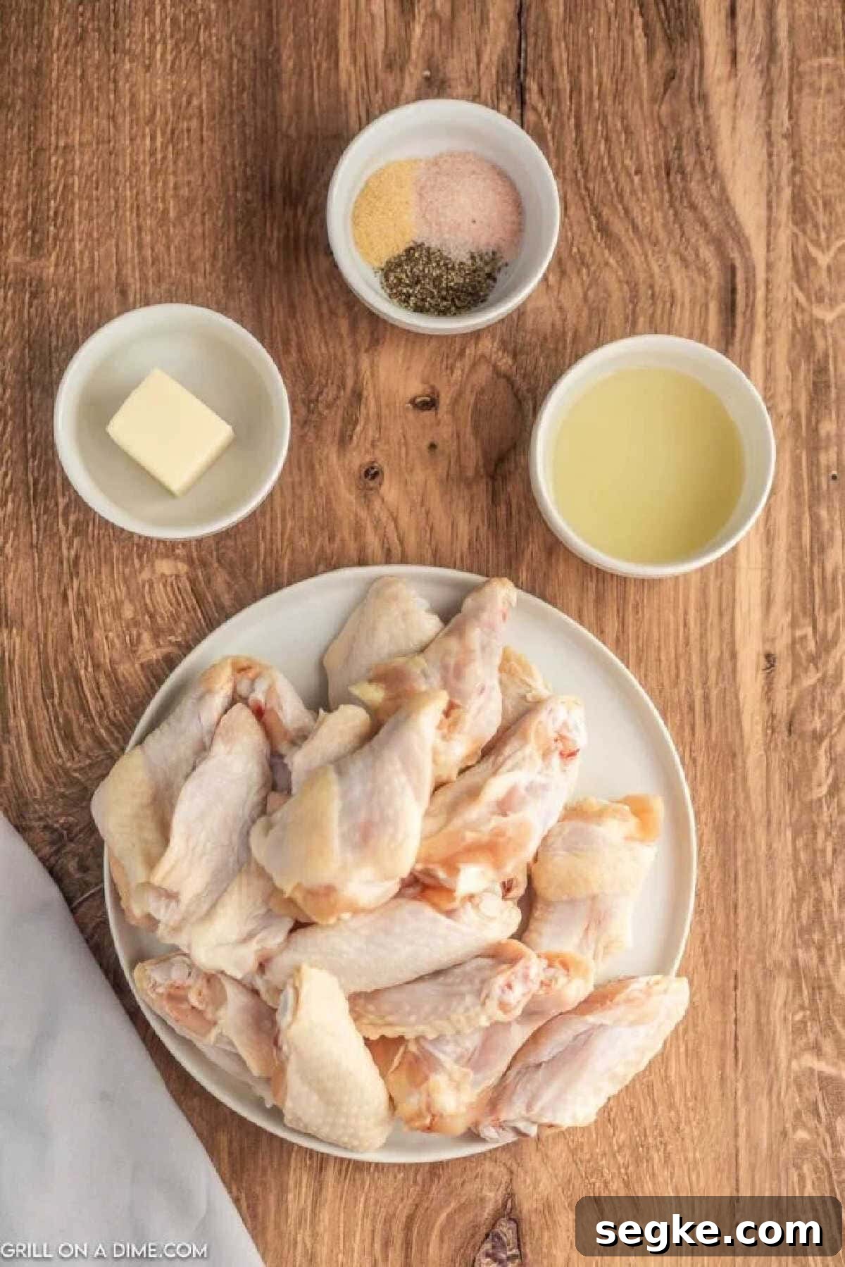Uncooked chicken wings stacked on a plate with a bowl of oil, butter, and salt, pepper and garlic powder in a bowl.